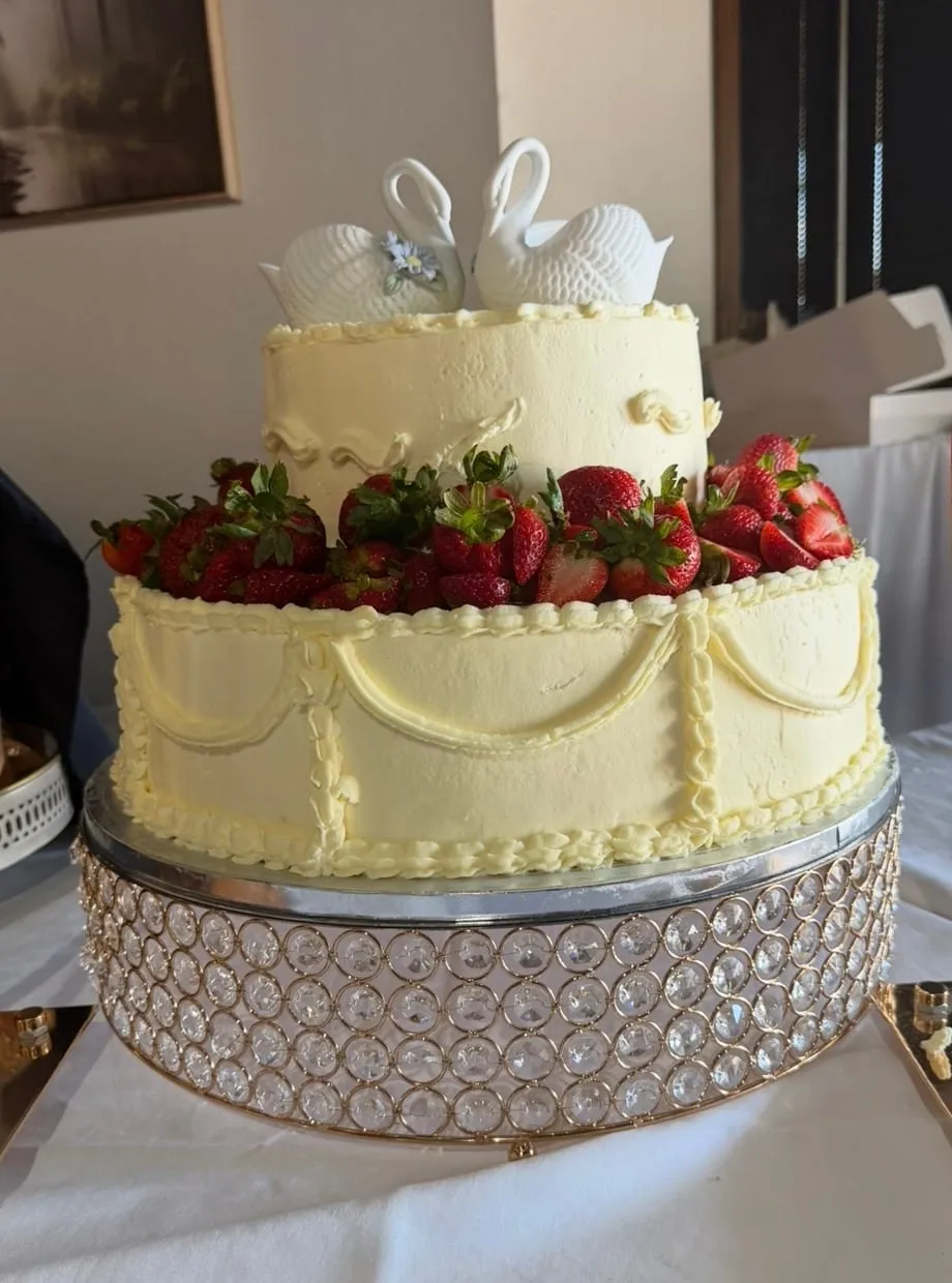 Two-tier white buttercream cake topped with fresh strawberries and a bow, on a rhinestone stand