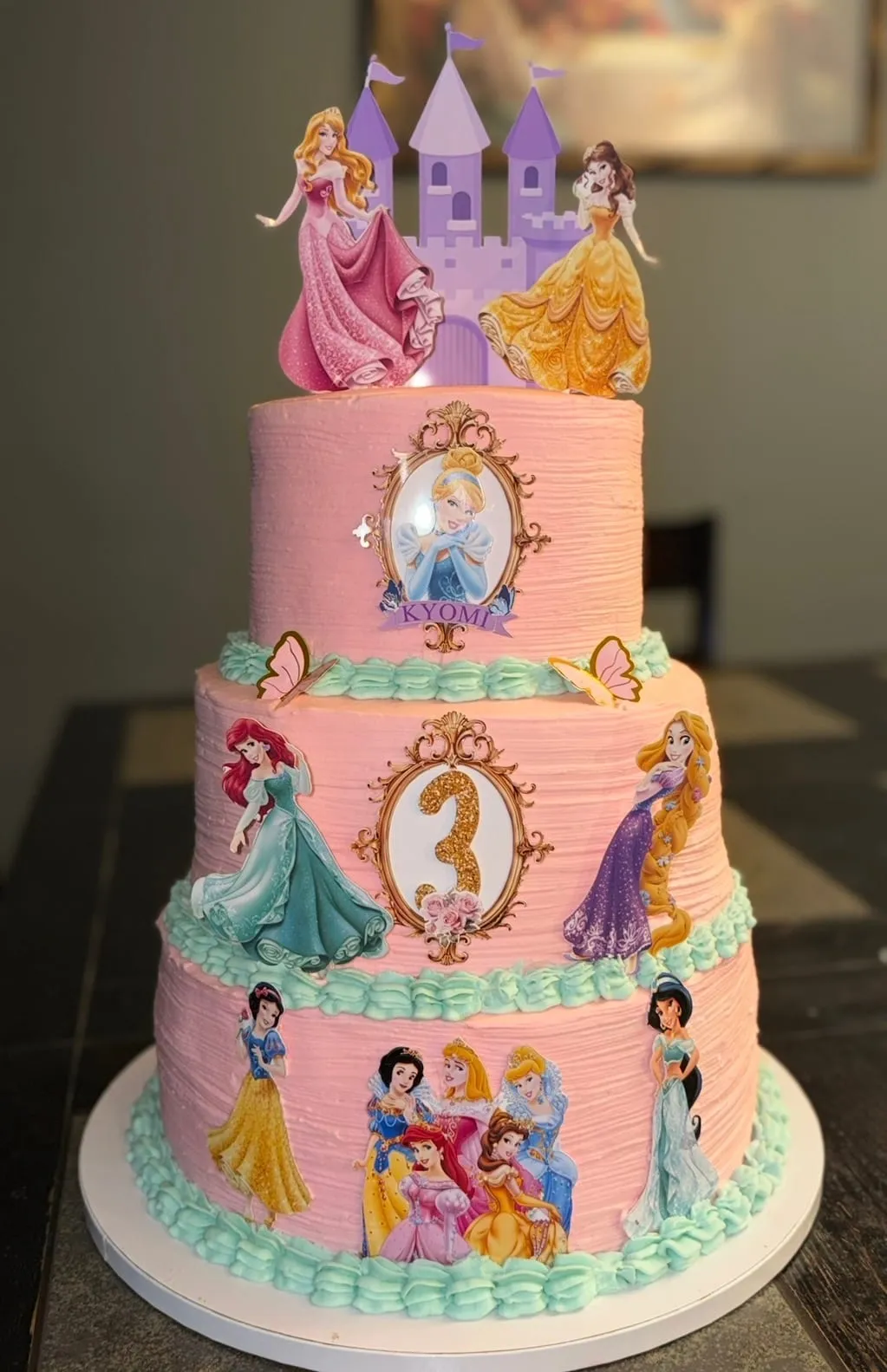 Three-tier pink cake with Disney princess figures and a gold number three, finished in soft pastel buttercream