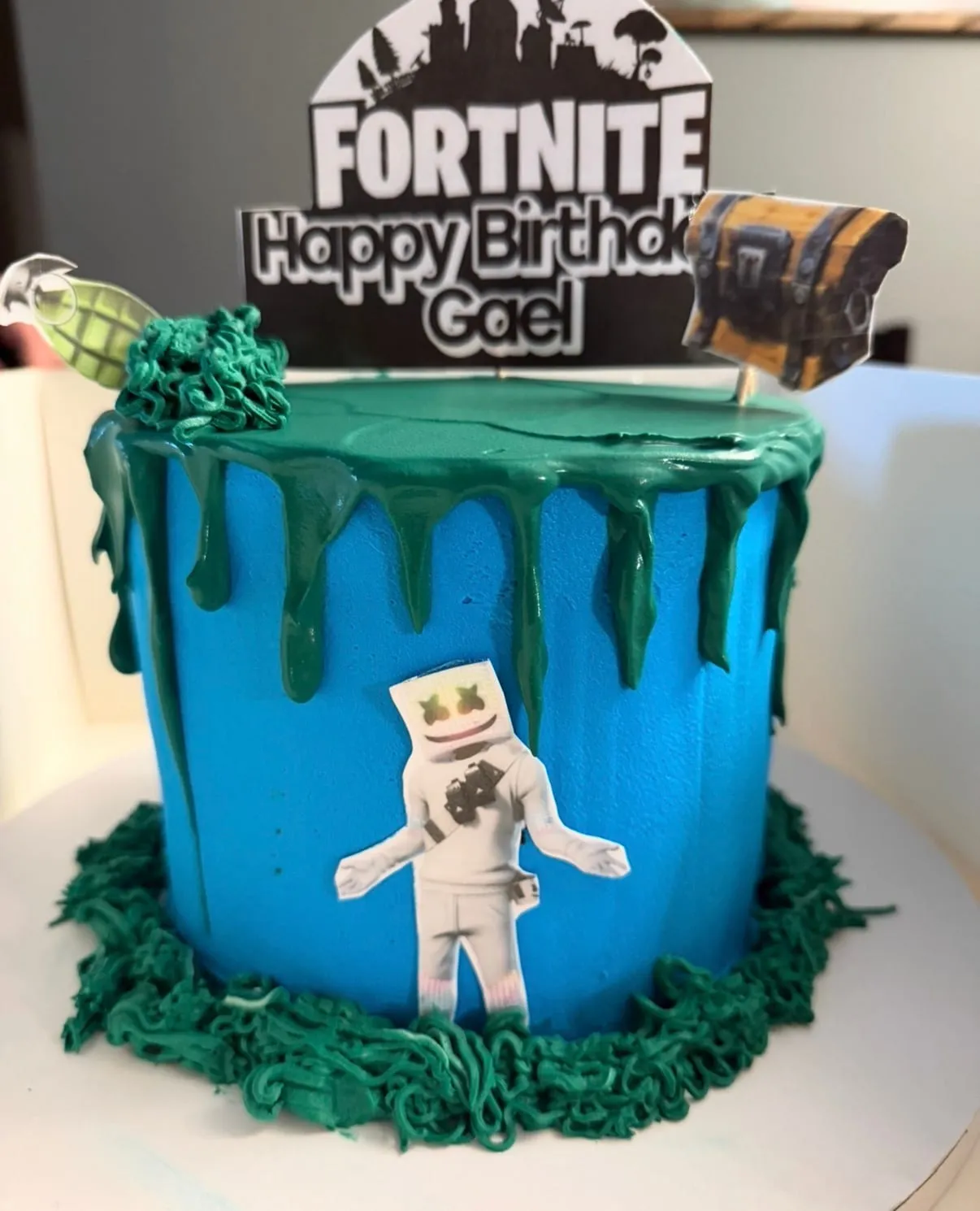 Blue drip cake with a Marshmello figure and green grass piping, topped with a Fortnite name banner