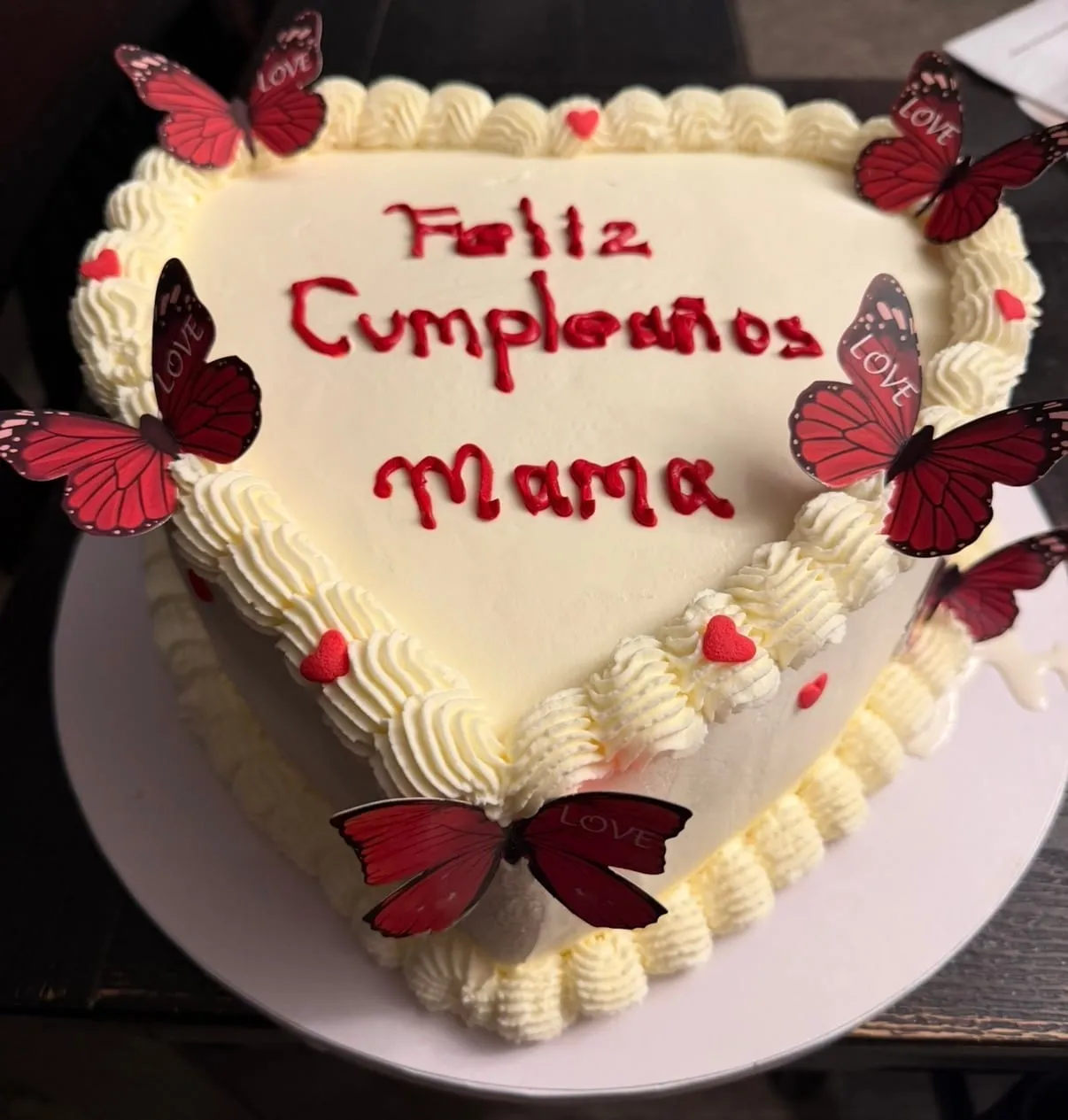 Cream heart-shaped cake with red script lettering, hearts, and red butterflies for 'Mamá'