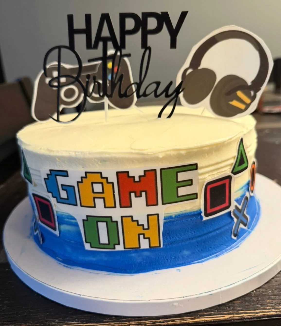 Gamer cake with PlayStation-button lettering spelling 'Game On' and a headphones-and-controller topper