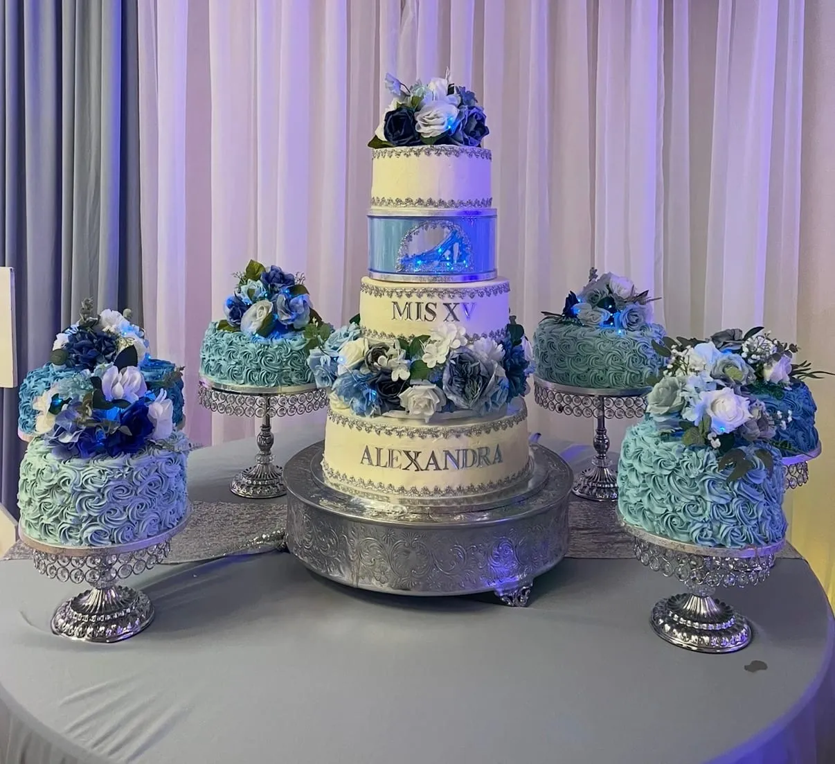 Three-tier quinceañera cake in powder blue and silver with sugar flowers and a name plaque, on a decorated table